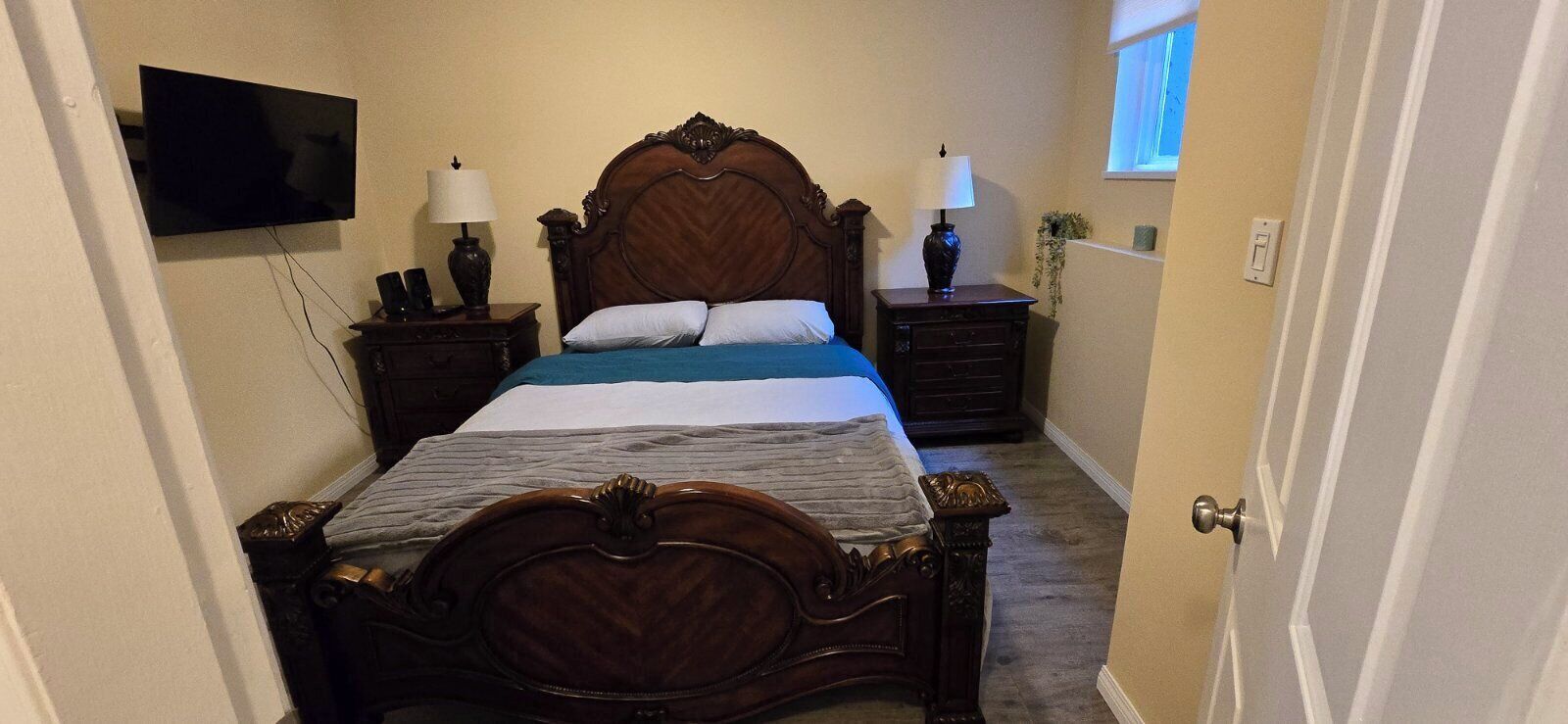 A narrow hotel room with a wooden bed, two bedside tables, a TV mounted on the wall, and a small window.