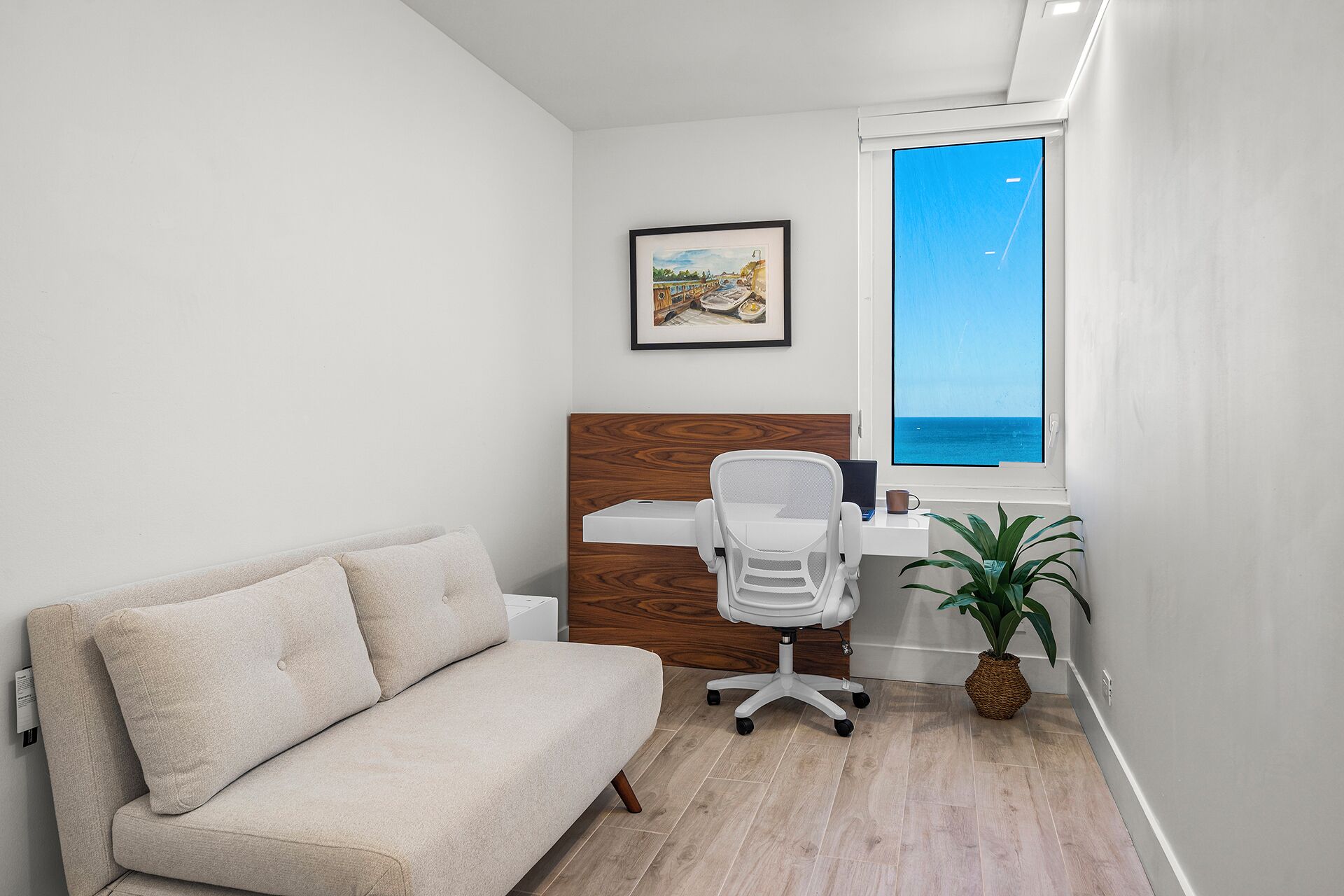 A modern, compact room with a white sofa, a wooden accent wall, a desk with a laptop, and a view of the sea through a window.