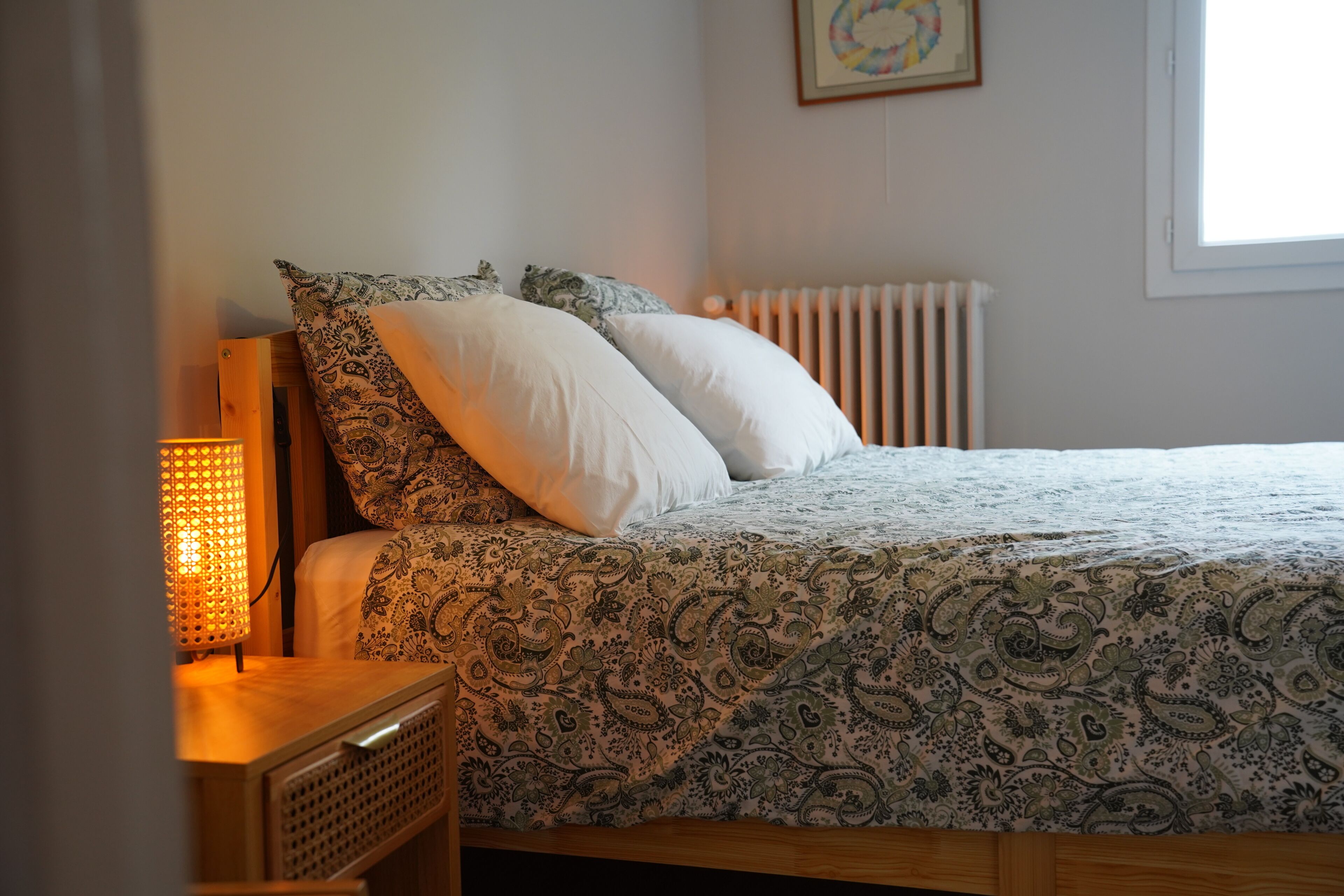 A bed with a patterned bedspread, two pillows, a bedside lamp, and a radiator in a room with a window.