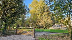 A gravel pathway leading to a wooden gate, surrounded by trees and a stone structure in the background.