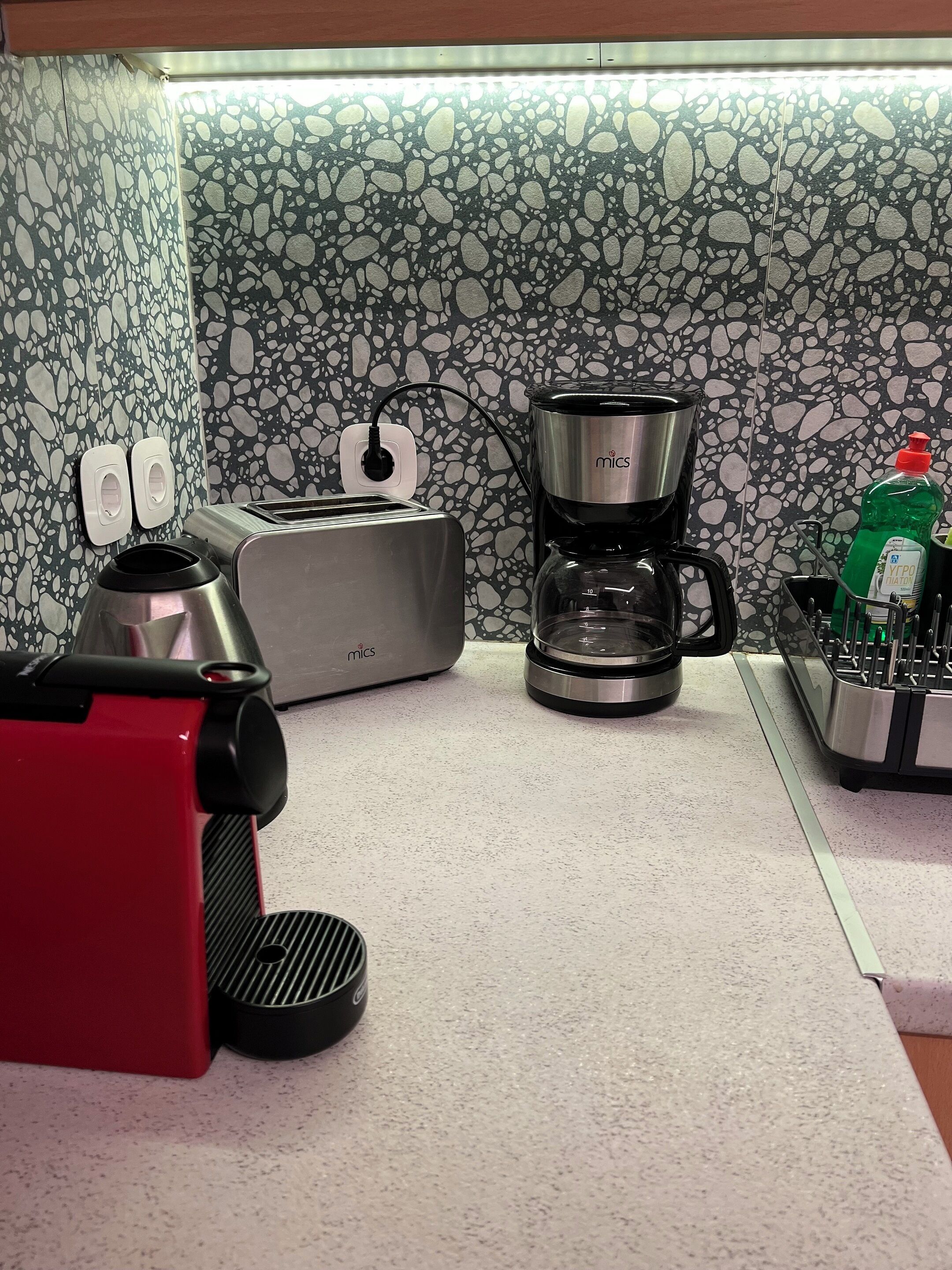 Una kitchenette con cafetera, tostadora y un electrodoméstico rojo, posiblemente una olla arrocera, frente a un salpicadero moteado.