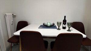 Ein Essbereich mit einem weißen Tisch, vier braunen Stühlen, einer Weinflasche aus Glas, zwei Weingläsern und einem dekorativen Teller.