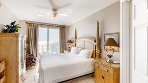 Une chambre d’hôtel avec un grand lit, un ventilateur de plafond, un balcon avec des rideaux, une commode en bois et une lampe.
