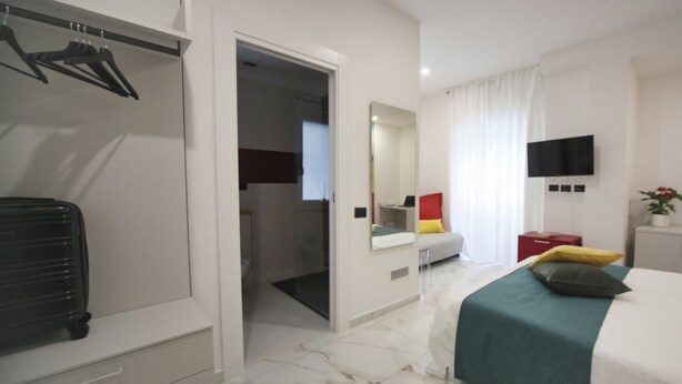 A hotel room with a bed, a wardrobe, a mirror, a television, and a bathroom visible through an open door. - Becoloured Circles Suite. Where it is possible to dream among the masterpieces of art. (Napoli)