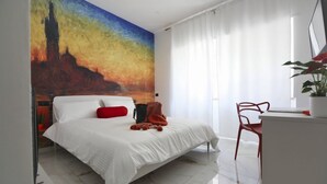 A hotel room with a bed, a desk, a chair, a large painting on the wall, and a window with curtains. - Venice Suite. Becoloured, where it is possible to dream among art masterpieces. (Napoli)