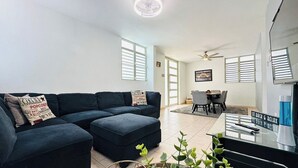 Un salon moderne avec un canapé d’angle bleu foncé, un coin repas comprenant une table et des chaises, et un ventilateur de plafond.