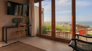 A room with a TV on a wall, a desk with a chair, a large sliding glass door leading to an outdoor area with a view of the sea. - Casa Kahu by Wynwood House (Los Organos)