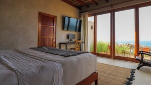A bedroom with a bed, a TV mounted on the wall, a desk with a chair, and a view of the ocean through large sliding doors. - Casa Kahu by Wynwood House (Los Organos)