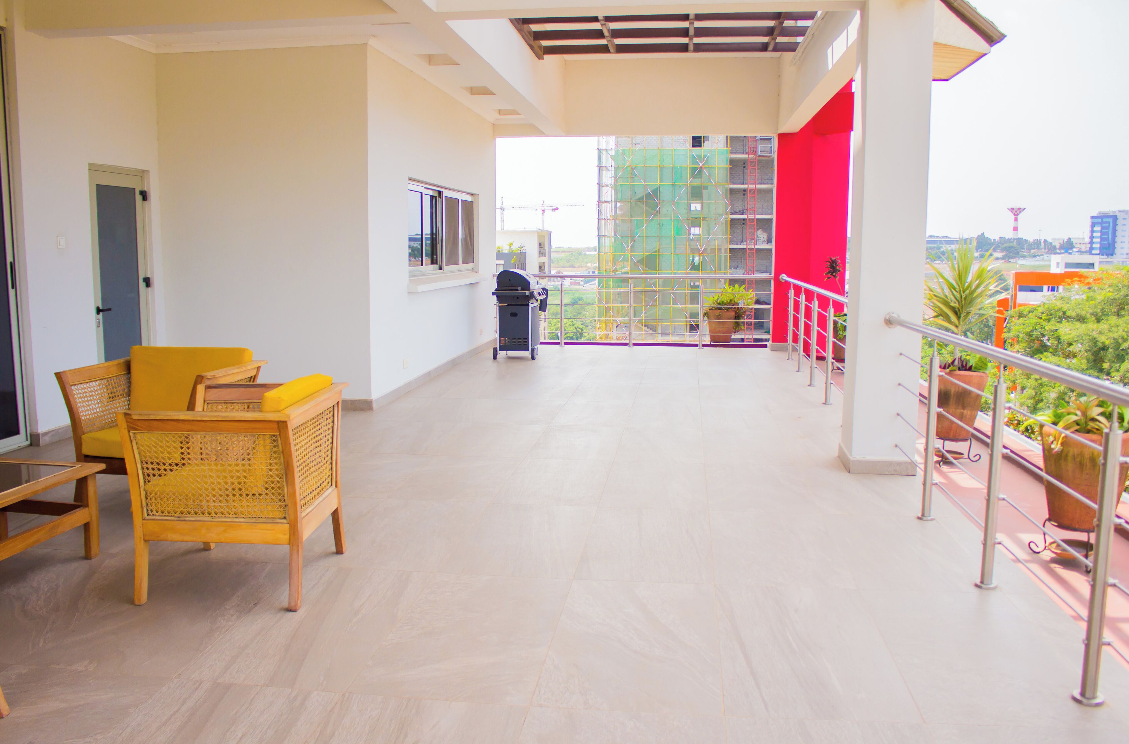 A spacious balcony with a wooden floor, yellow chairs, a grill, and potted plants.