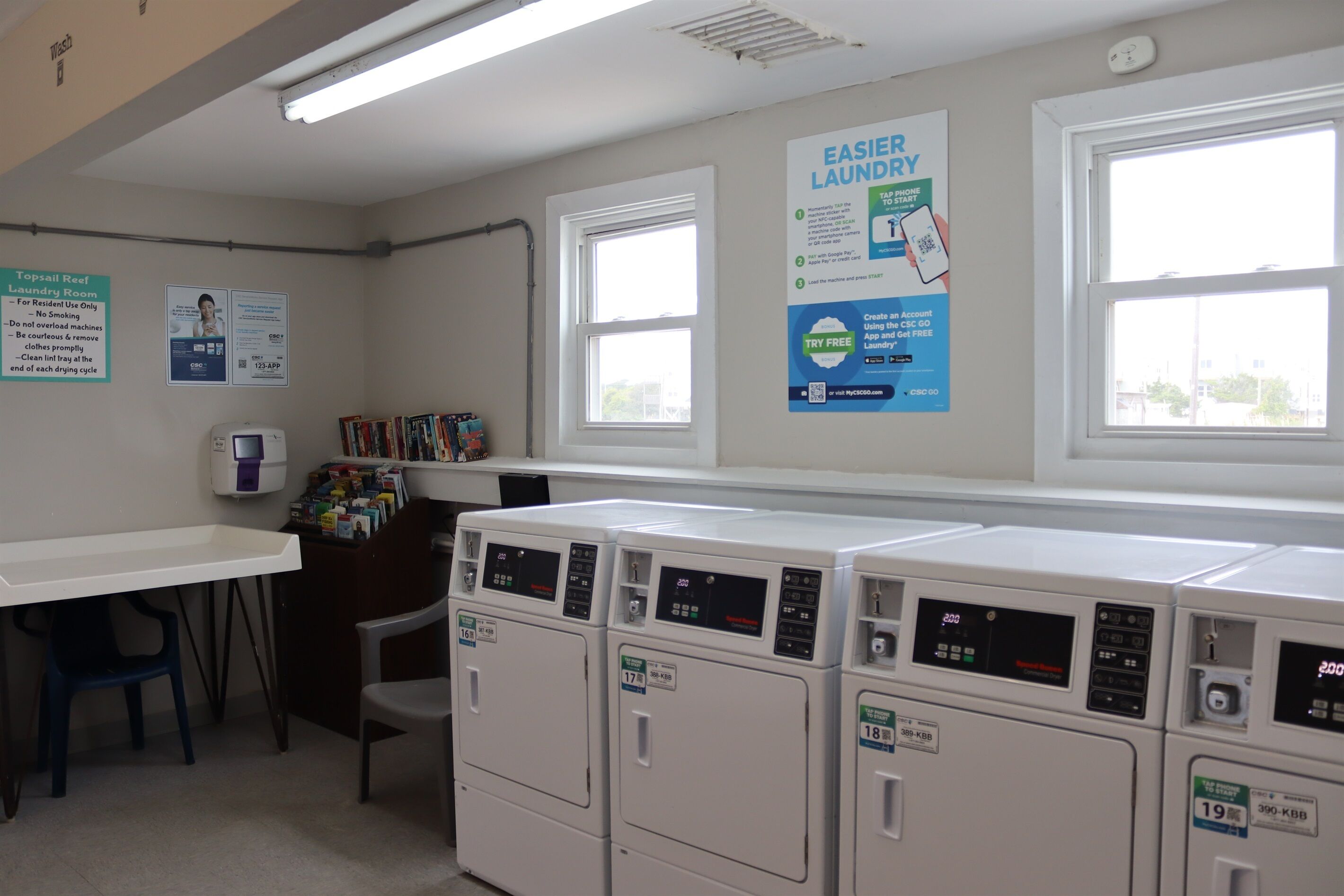 Una sala de bugaderia amb diverses rentadores, una taula, cadires i cartells informatius a les parets.