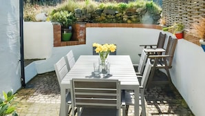 An outdoor patio with a white table and chairs, a vase of yellow flowers, and a stone wall with plants.