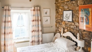 A bedroom with a stone wall, a bed with white bedding, a window with curtains, and framed artwork on the wall.