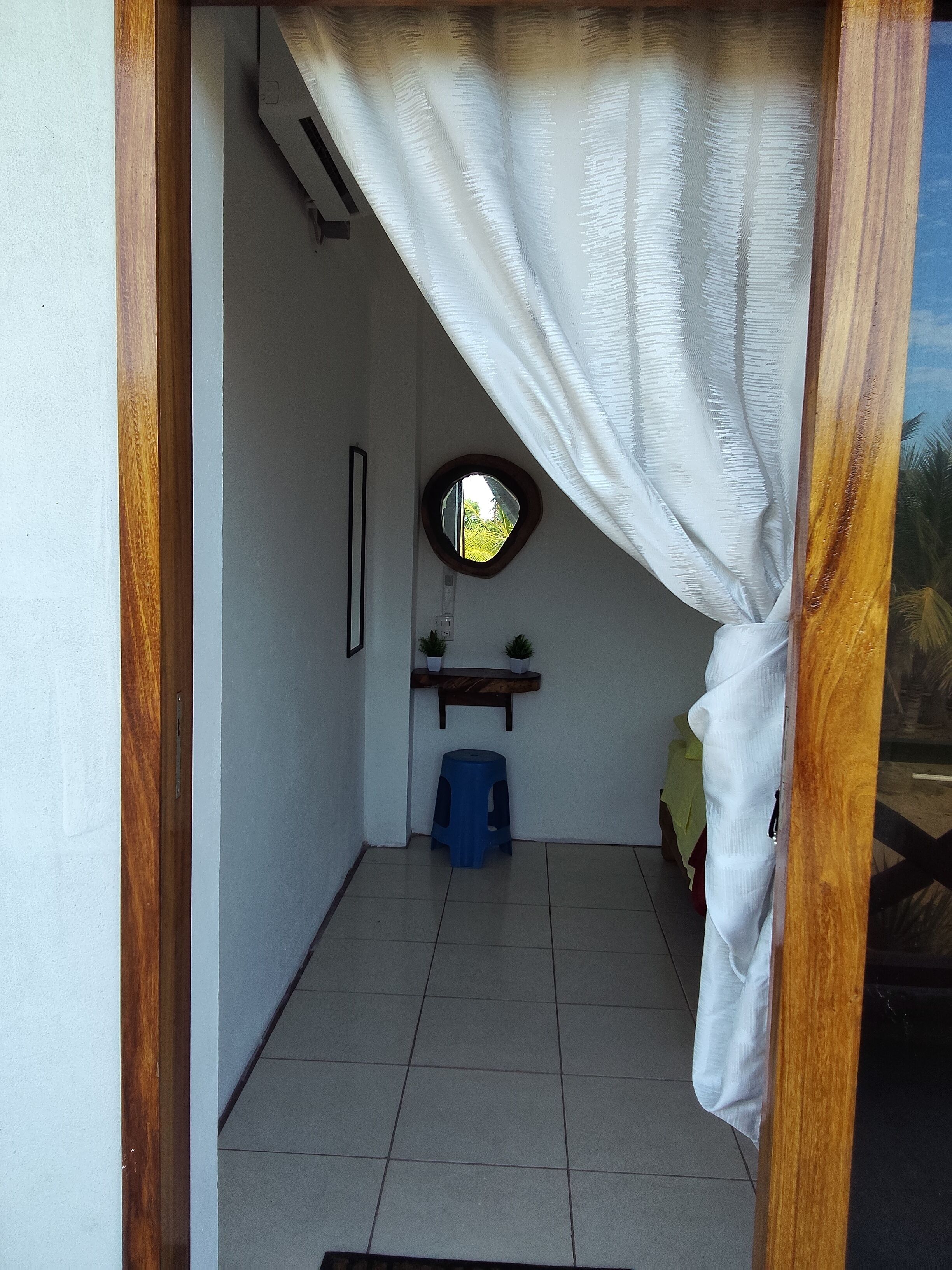 A narrow hallway with a wooden door frame, a small stool, a shelf with two potted plants, and a round mirror on the wall.
