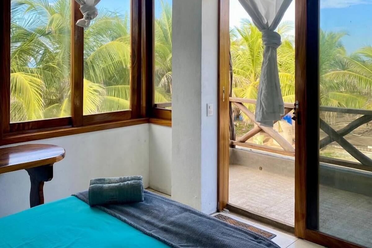 A room with a bed, a small table, and a view of palm trees through large windows.