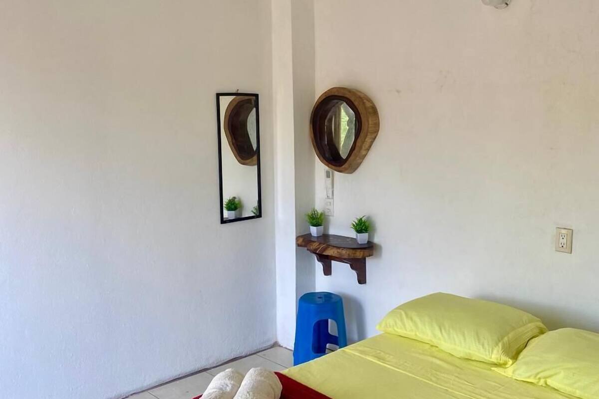 A hotel room with a bed, two wall-mounted mirrors, a small wooden shelf with plants, a blue stool, and a red blanket.