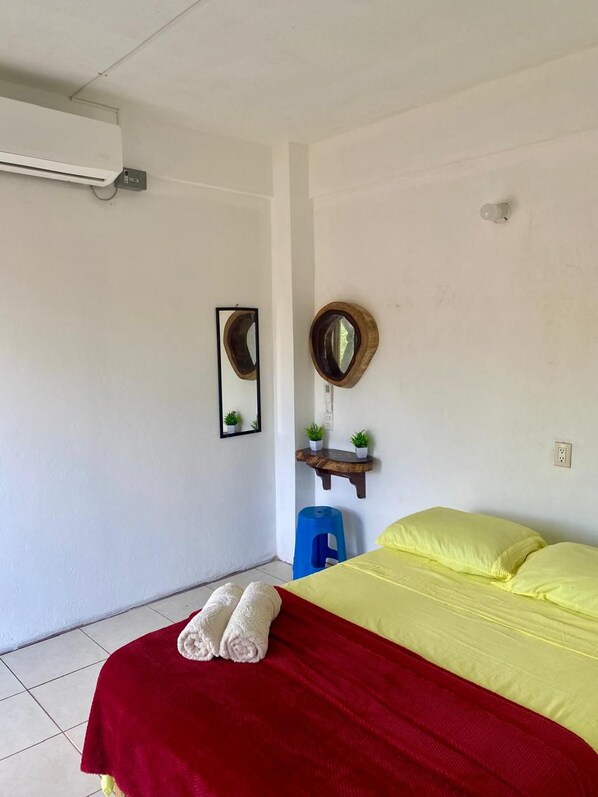 A hotel room with a bed, two wall-mounted mirrors, a small wooden shelf with plants, a blue stool, and a red blanket.