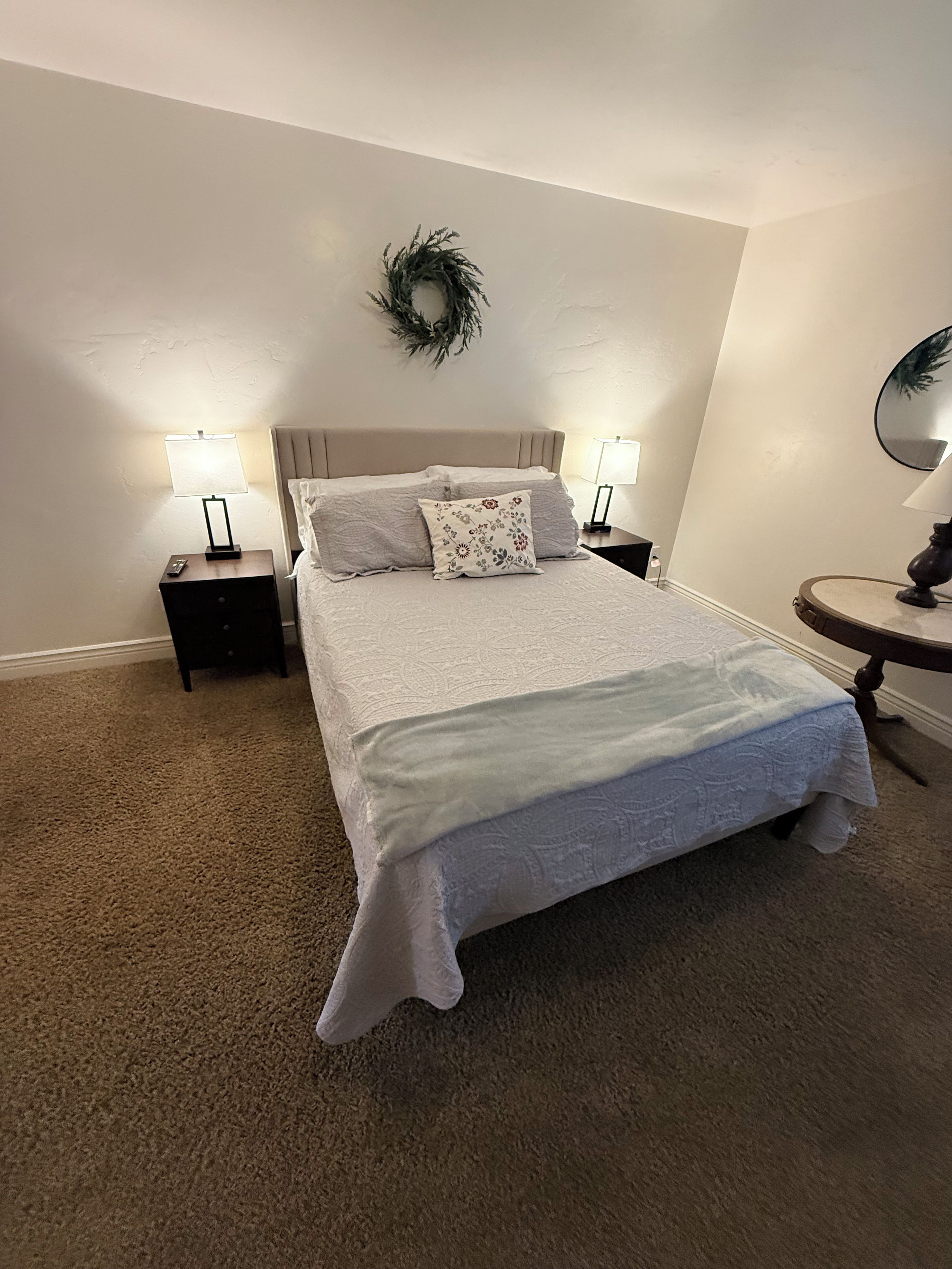 Une chambre à coucher avec un grand lit, deux tables de chevet avec des lampes, un miroir rond et une couronne décorative accrochée au mur.