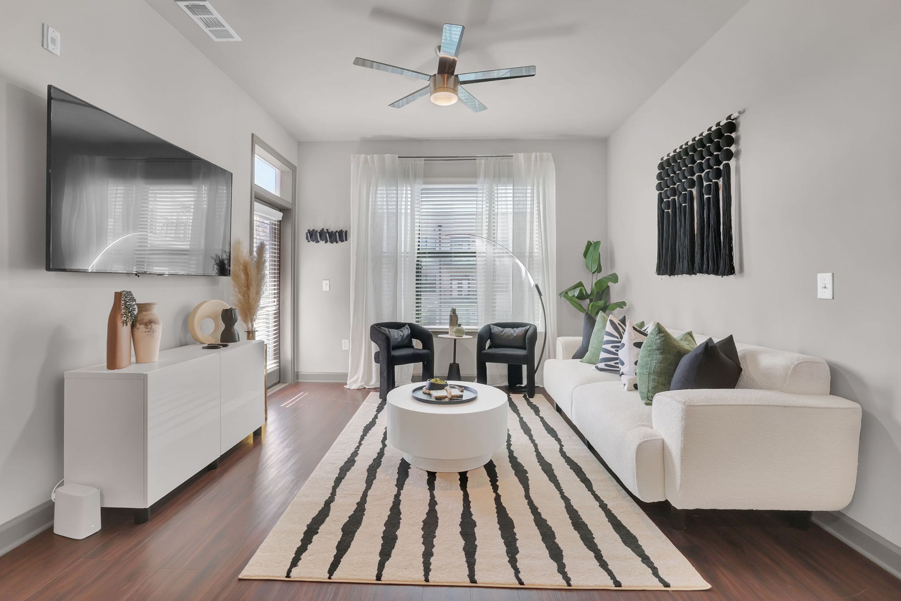 A modern living room with a white sofa, a coffee table, and a TV mounted on the wall. There is a ceiling fan, a window with blinds, and a decorative wall piece.