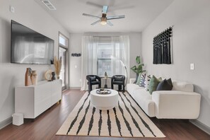 A modern living room with a white sofa, a coffee table, and a TV mounted on the wall. There is a ceiling fan, a window with blinds, and a decorative wall piece. - Ebony Ivory Midtown King Bed (Atlanta)
