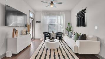 A modern living room with a white sofa, a coffee table, and a TV mounted on the wall. There is a ceiling fan, a window with blinds, and a decorative wall piece.