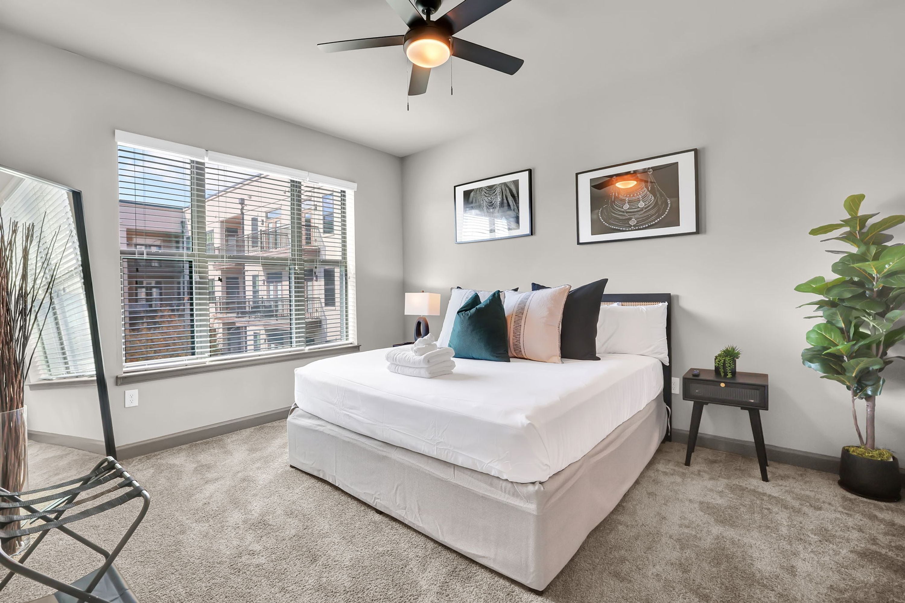 A bedroom with a large bed, a ceiling fan, a window with blinds, and a small bedside table with a plant.