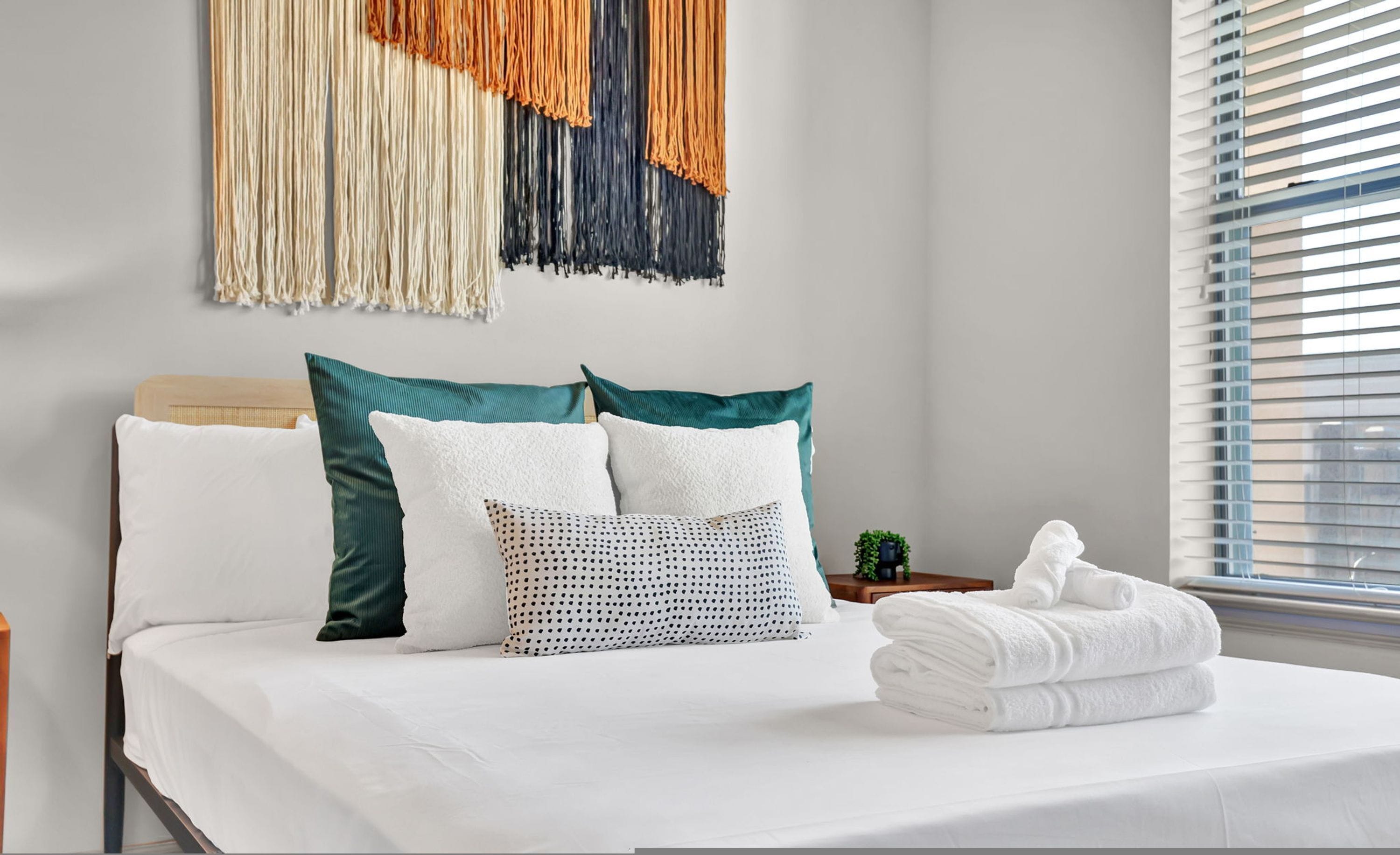 A neatly made bed with white linens and a variety of pillows, including a polka dot one. A small potted plant is on the nightstand beside the bed.