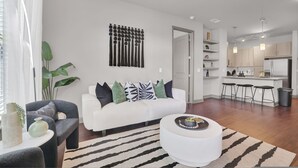 Un salón moderno con un sofá blanco, un sillón gris y una mesa redonda de centro. Se ve una zona de cocina con una barra y taburetes a través de una puerta abierta.