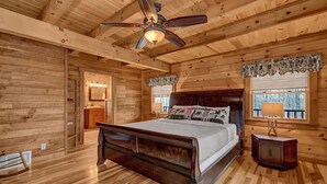 A wooden cabin bedroom with a bed, a nightstand, a ceiling fan, and a window with curtains. - Treehouse Lodge: Luxury Log Cabin Retreat in Hocking Hills (Laurelville)