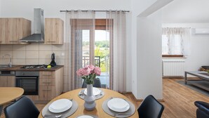 Eine moderne Küche mit Gasherd, Backofen und Holzschränken. Ein Essbereich mit rundem Tisch für vier Personen, und Blick in einen Garten durch ein Fenster mit Spitzenvorhängen.