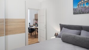 Ein Schlafzimmer mit einem Bett, einem Schreibtisch mit Stühlen und Blick auf ein historisches Gebäude durch eine offene Tür.