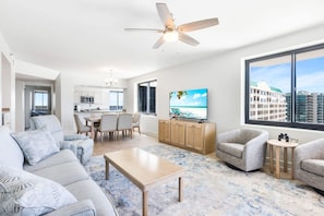 A modern living room with a sofa, chairs, a coffee table, and a TV. There is a ceiling fan, a kitchen in the background, and a large window with a view of buildings.