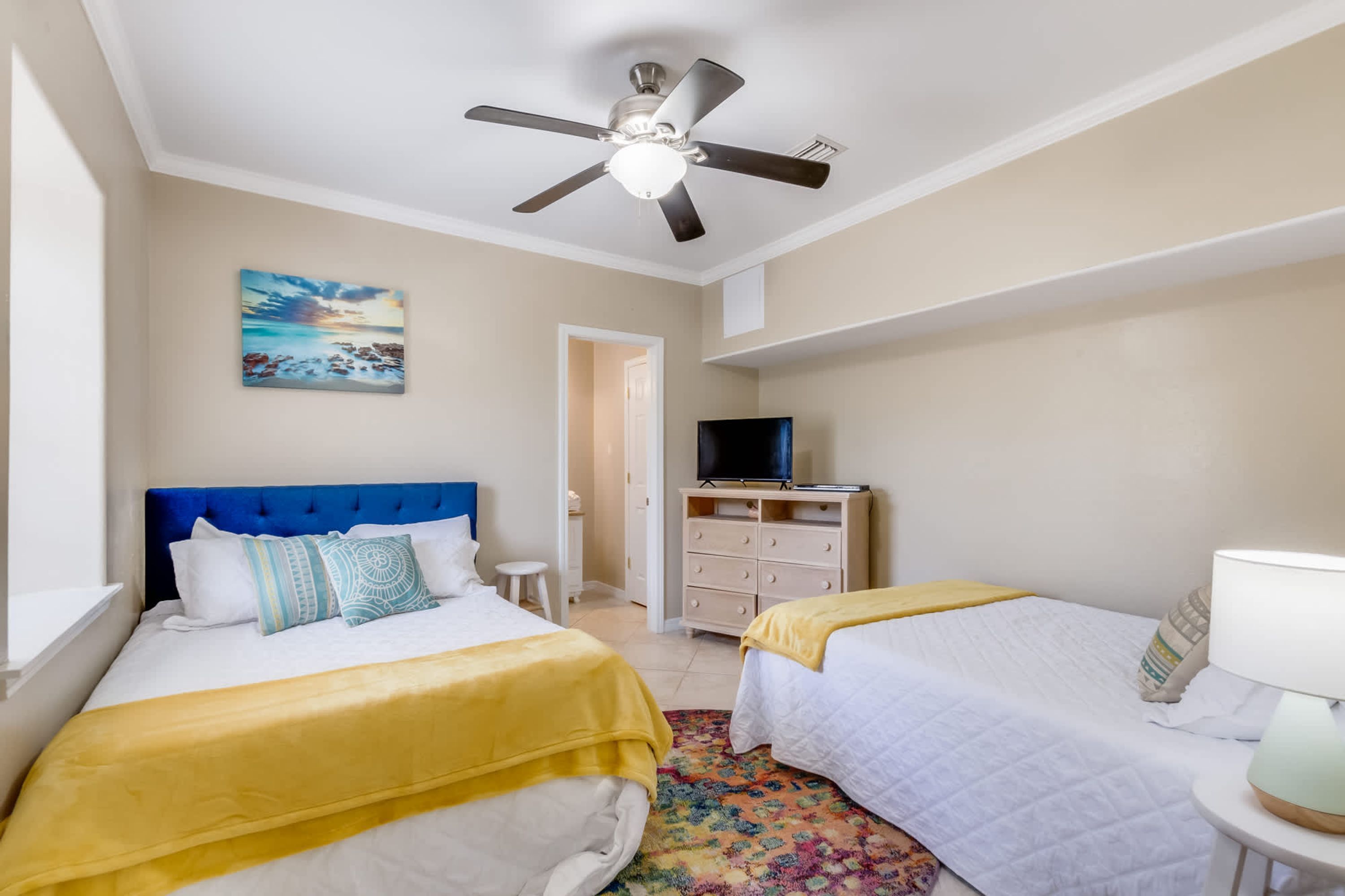 Habitación de hotel con dos camas, ventilador de techo, televisor y un cuadro en la pared.