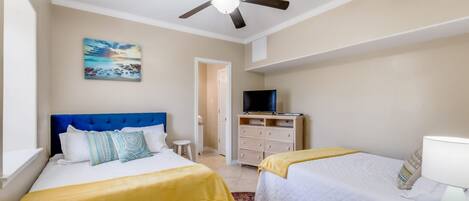 Habitación de hotel con dos camas, ventilador de techo, televisor y un cuadro en la pared.