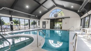 En indendørs swimmingpool med klart vand, omgivet af hvide stole og borde, store vinduer og et hvælvet loft med synlige bjælker.