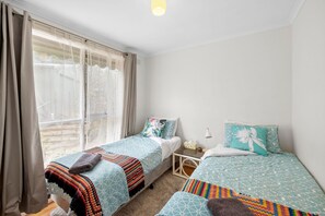 Ein Schlafzimmer mit zwei Betten, einem Fenster mit Vorhängen, einem Nachttisch mit Lampe und Blick ins Freie.