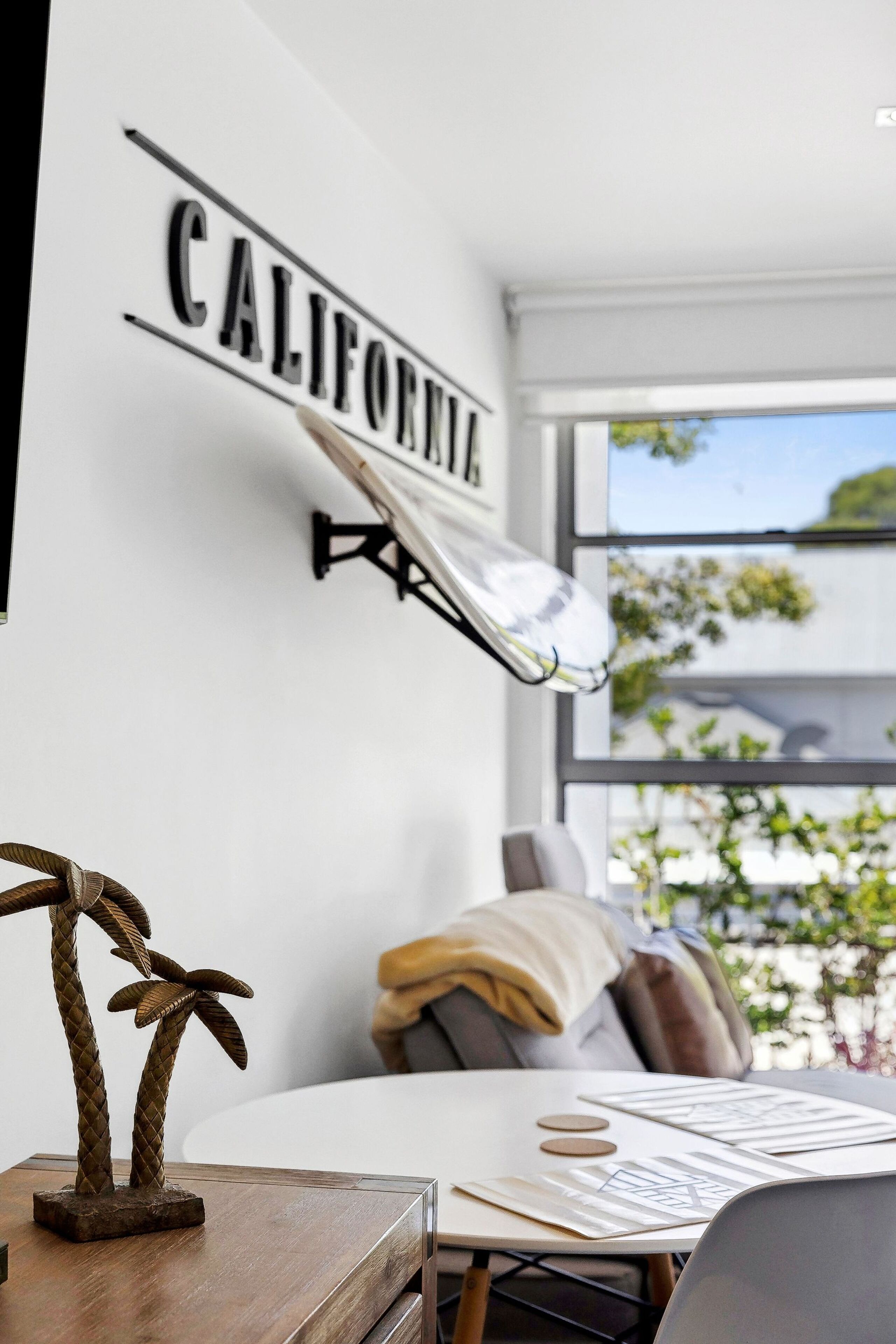 Een kamer met een wandlamp, een houten tafel met een decoratief object, en een raam met uitzicht op groen.
