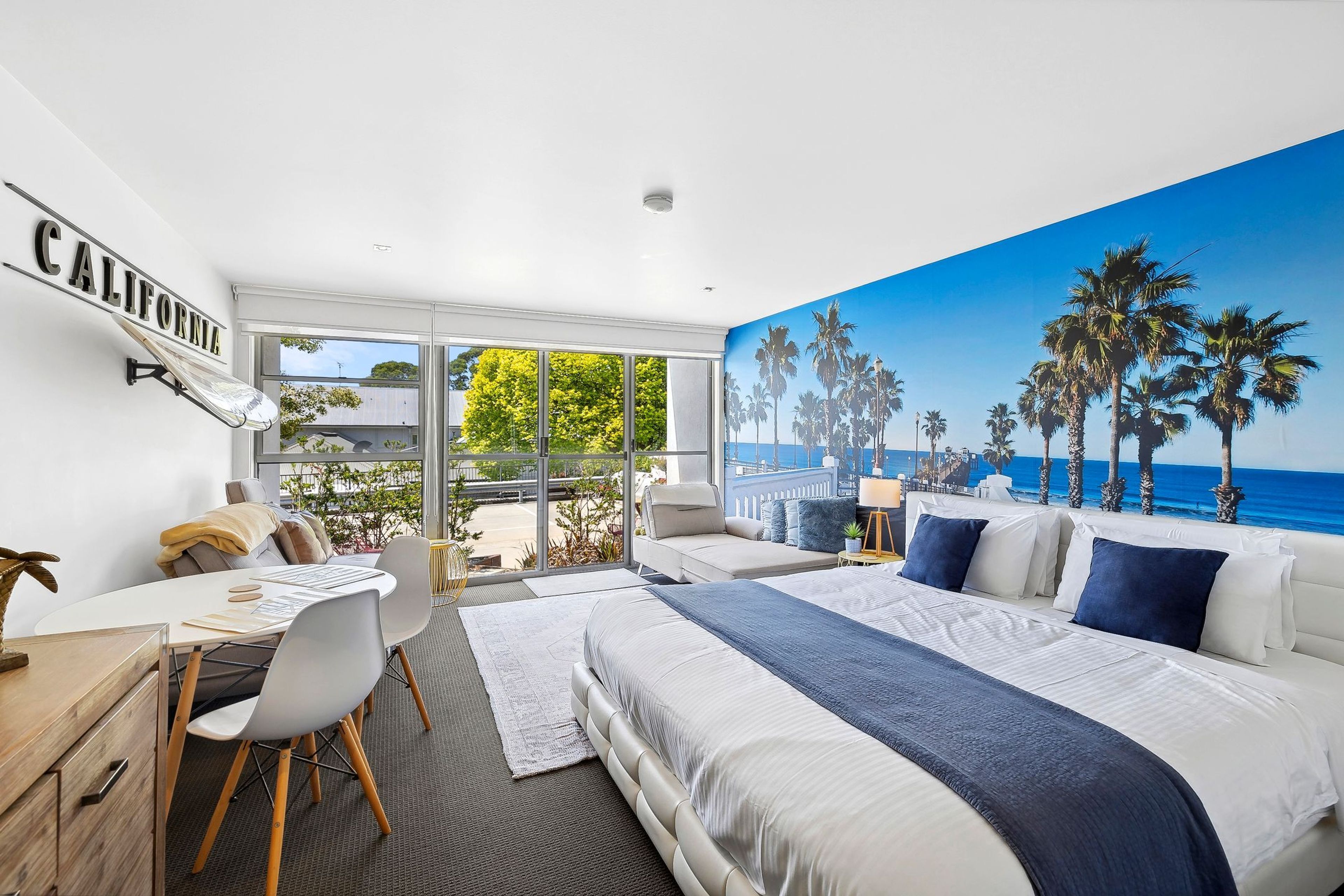 A hotel room with a bed, a desk, and a chair. There is a large window with a view of palm trees and the ocean.