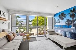 A modern living room with a large sliding glass door, a white sofa, and a view of a residential area. - Bay View Motel - California Beach (Lorne)