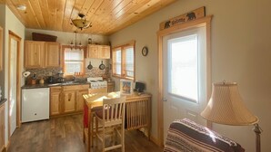 Interior acogedor de una cabaña con piso de madera, zona de comedor, cocina con gabinetes de madera, una silla roja y una lámpara.