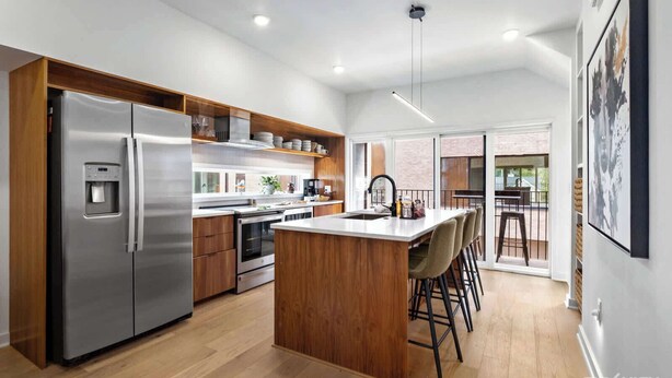 Une cuisine moderne dotée d’un réfrigérateur en acier inoxydable, de meubles en bois, d’un îlot central avec des tabourets de bar et d’une grande fenêtre donnant sur un balcon.