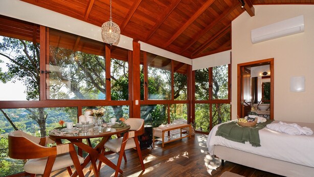 A spacious room with a large window, a bed, a dining table with chairs, and a bench. - Charmosa Suíte em Sambaqui - NDI005 (Florianópolis)