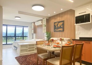 Una habitación de hotel moderna con una cama, un área de comedor con una mesa preparada para dos personas, una cocina con microondas y un ventanal con vistas al exterior.