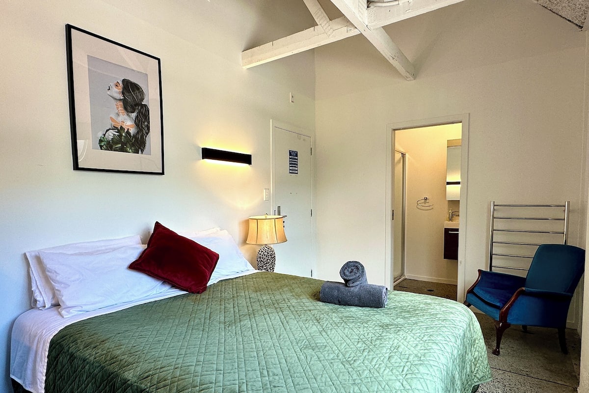 A hotel room with a bed, a red pillow, a green quilted bedspread, a wall-mounted lamp, a towel, a chair, and a door leading to a bathroom.