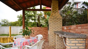 Un patio al aire libre con una parrilla de ladrillo, sillas de plástico blanco y una mesa con mantel floreado.