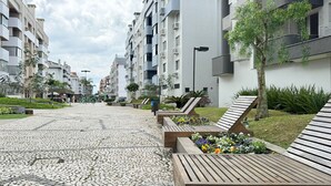 A paved walkway with benches and flower beds in a residential area with modern buildings. - Apartamento no Jurerê Open Shopping - NDI007 (Florianópolis)
