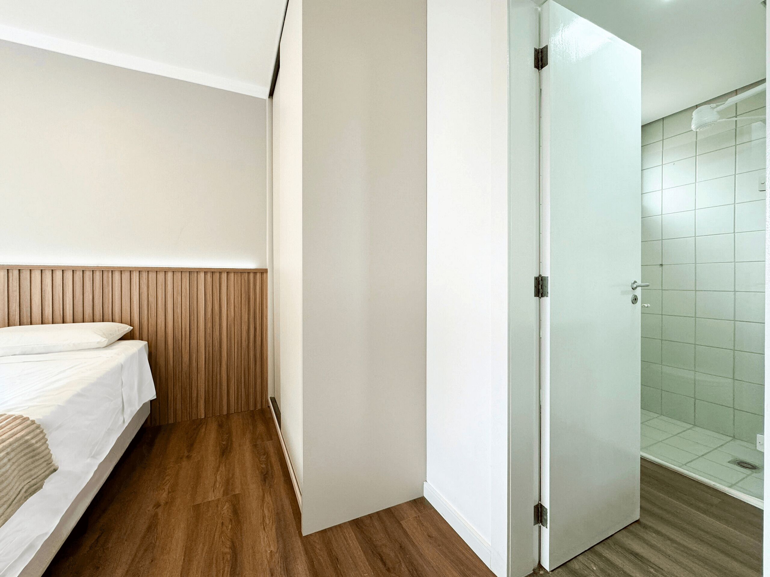 Une chambre à coucher avec une tête de lit en bois, un lit avec une literie blanche et une porte blanche donnant sur une salle de bain équipée d’une douche.