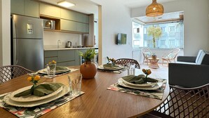 A modern kitchen with built-in appliances, a dining area with a wooden table set for four, and a view of the city through large windows. - Apartamento no Jurerê Open Shopping - NDI007 (Florianópolis)