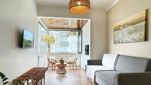 A modern living room with a grey sofa, wicker furniture, a hanging light fixture, and a painting on the wall. - Apartamento no Jurerê Open Shopping - NDI007 (Florianópolis)