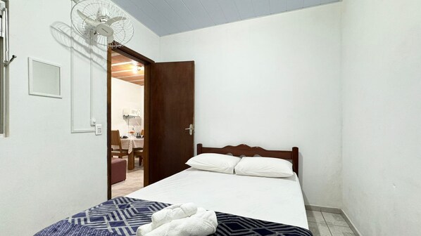Un dormitorio con cama, mesita de noche, ventilador y puerta que da a un baño.