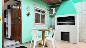 Una pequeña cocina con una mesa de madera y sillas blancas, una ventana con rejas de metal y una pared de azulejos con chimenea.